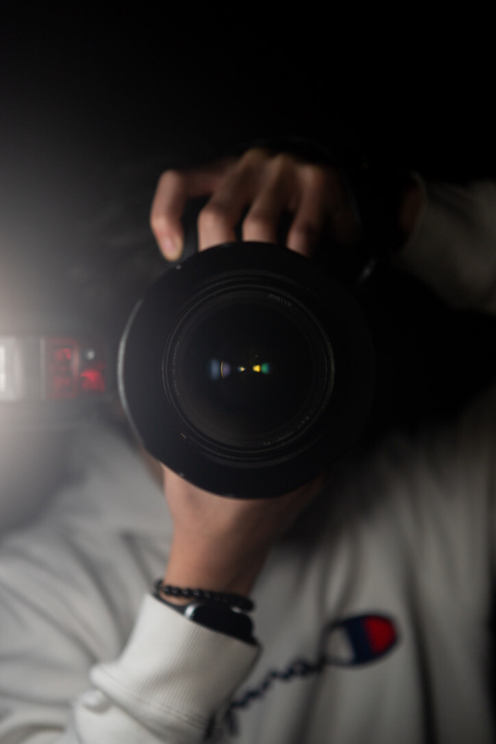 A photographer holding a large camera lens toward a mirror in a dim self-portrait