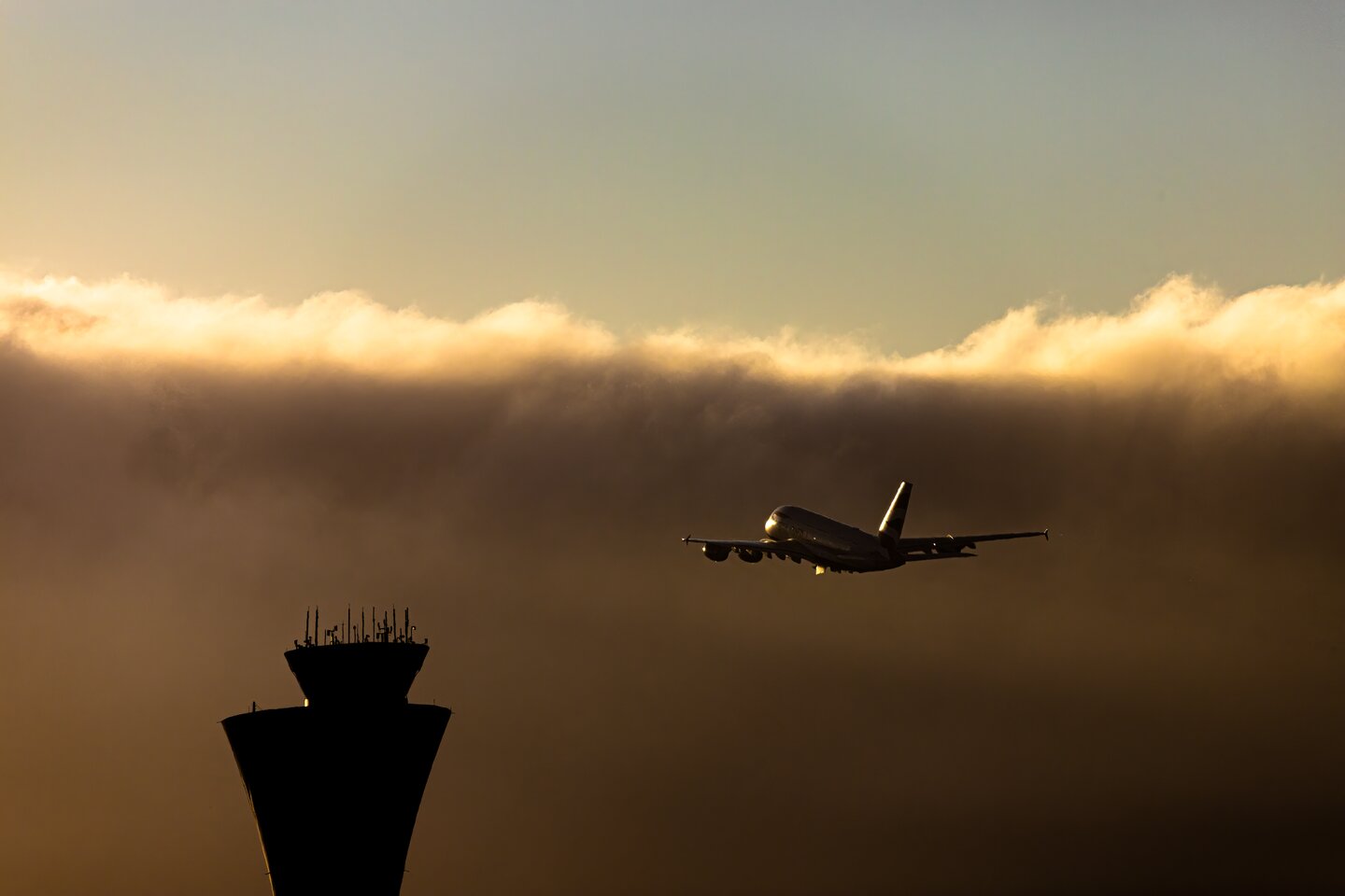 A departing aircraft silhouetted beneath gold clouds beside an airport control tower