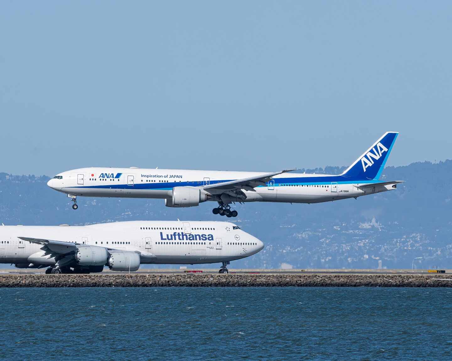 An ANA aircraft descending above a Lufthansa 747 beside San Francisco Bay
