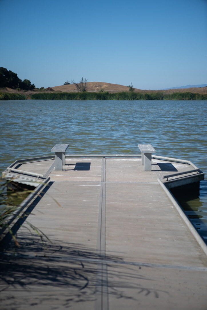A narrow dock with two seats extending into open water toward reeds and dry hills